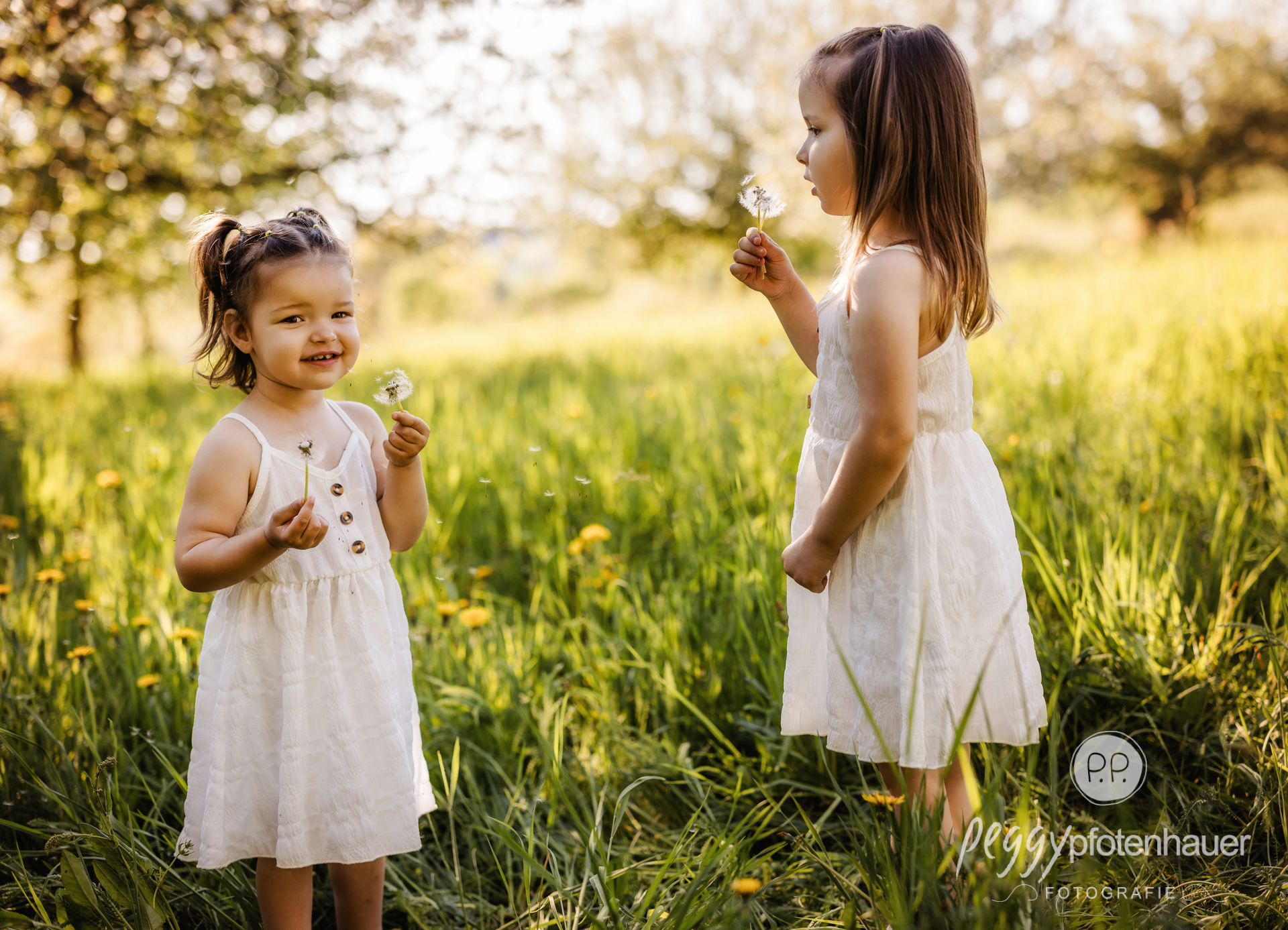 zwei Mädchen stehen in Frühlingswiese und pusten Pustblumen