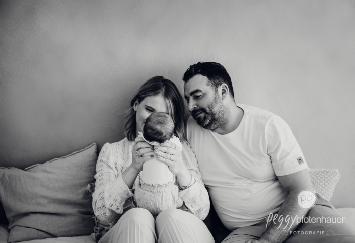 Mama und Papa sitzen auf Bett schauen sich Baby an