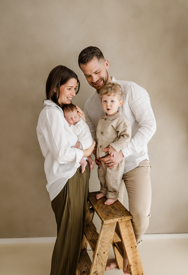 Familie steht zusammen mit Neugeborenem in Mamas Arm und Kleinkind in Papas Arm und lacht